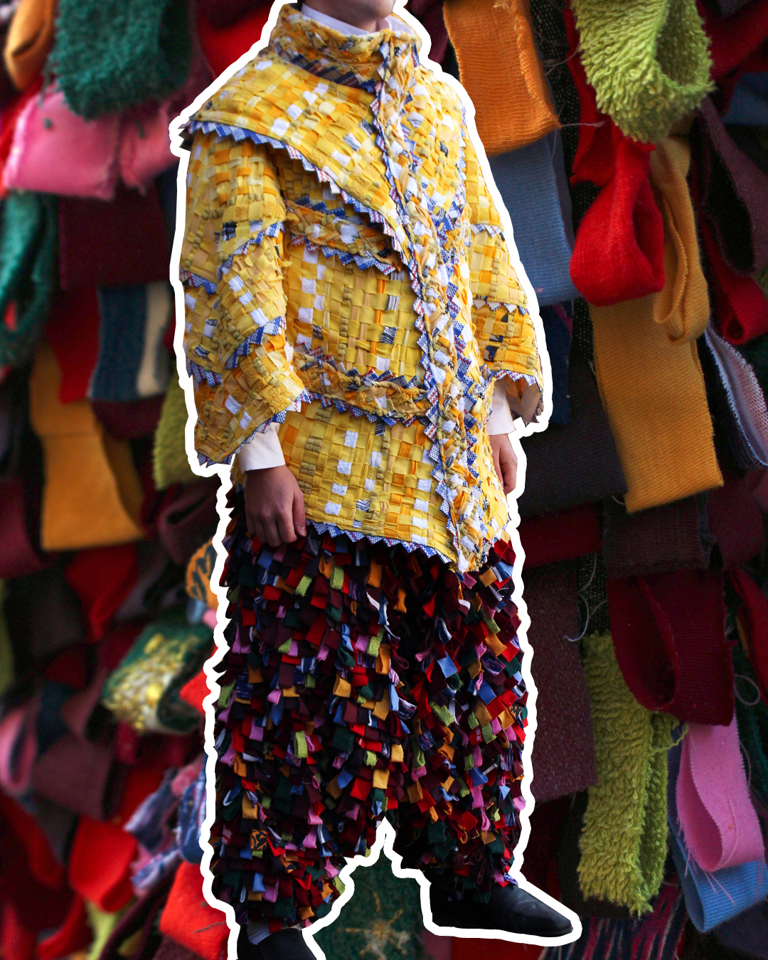 A photo of one of the characters made into a real outfit. A jacket made up of panels of yellow and white rags woven into a basket type surface. Poofy trousers made out of a myriad of short, colourful rag loops. The jacket has edge detailing made out of pale red and blue plastic, creating a zig-zag pattern.