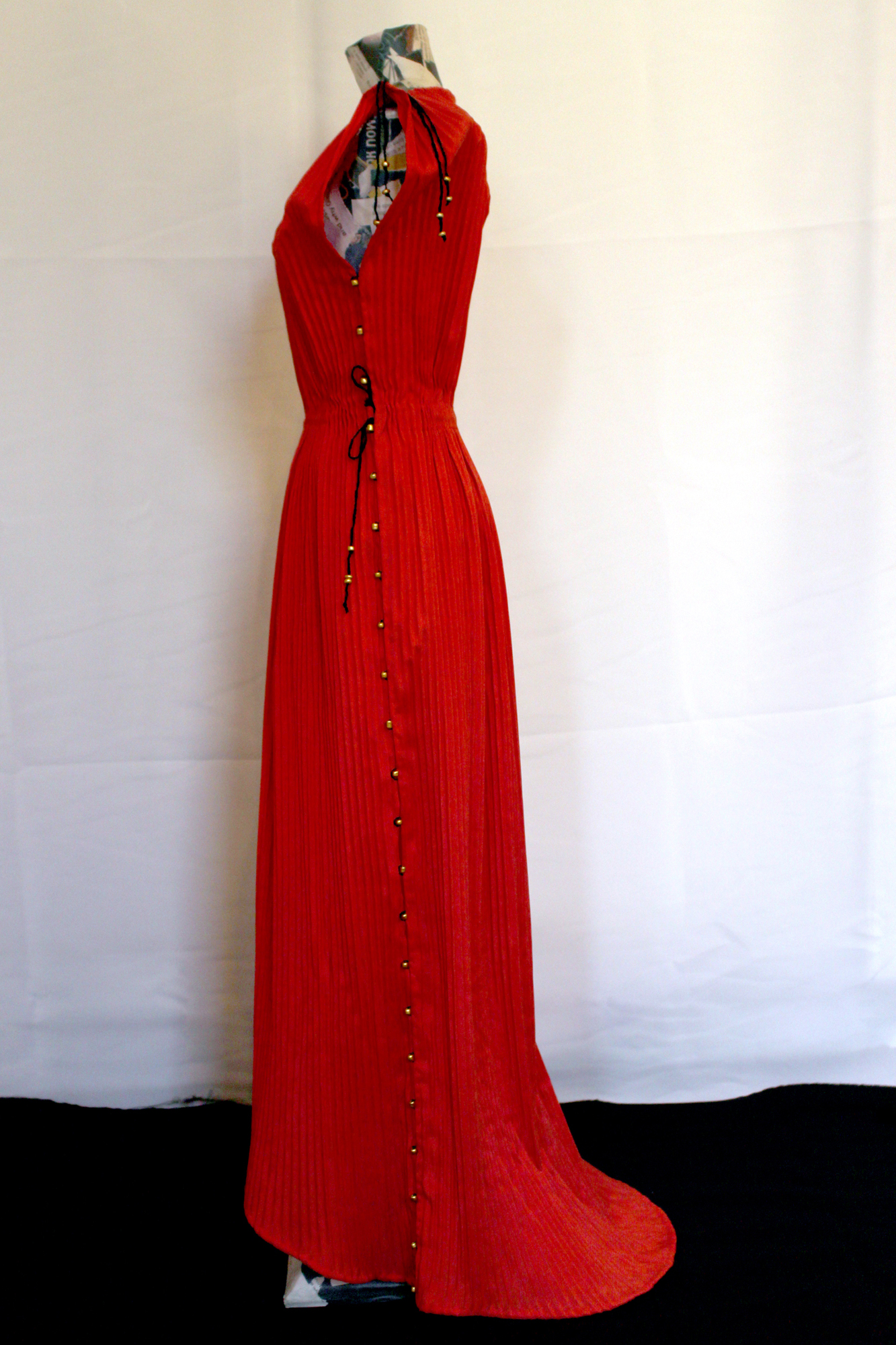 A full photo of the above dress, seen from the side and without the belt. This view shows the slight trail at the back of the dress, the black waist ties, and the full length row of golden bead closures along the side.