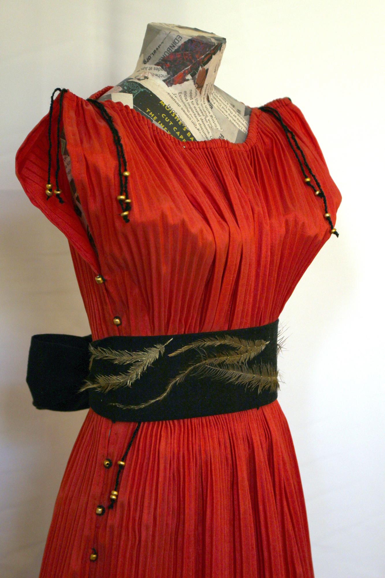 A waist-up photo of a red taffeta dress on a body form. The dress is narrowly pleated to conform to the shape of the body form. The shoulders have black closing ties finished with gold beads. The same gold beads go along the sides of the dress to button it closed. The waist has a wide black sash with gold emu feathers, tied into a bow at the back.