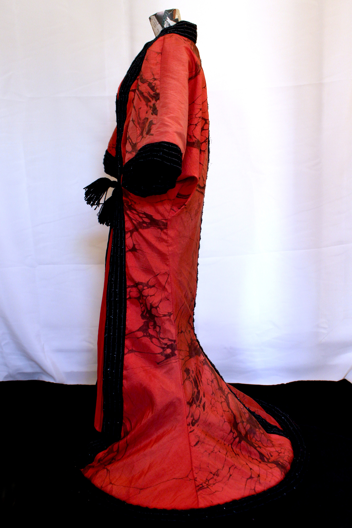 A full photo of the above dress, seen from the side, but with an opera coat over it. The coat is made from the same fabric, only covered in black marble detailing. The sleeves are wide and go to about elbow length. The black collar stands up at the neck and continues down the front. At the knees, the coat flares out towards the floor where it spreads into a circle shape. The waist is tied with black tassles.