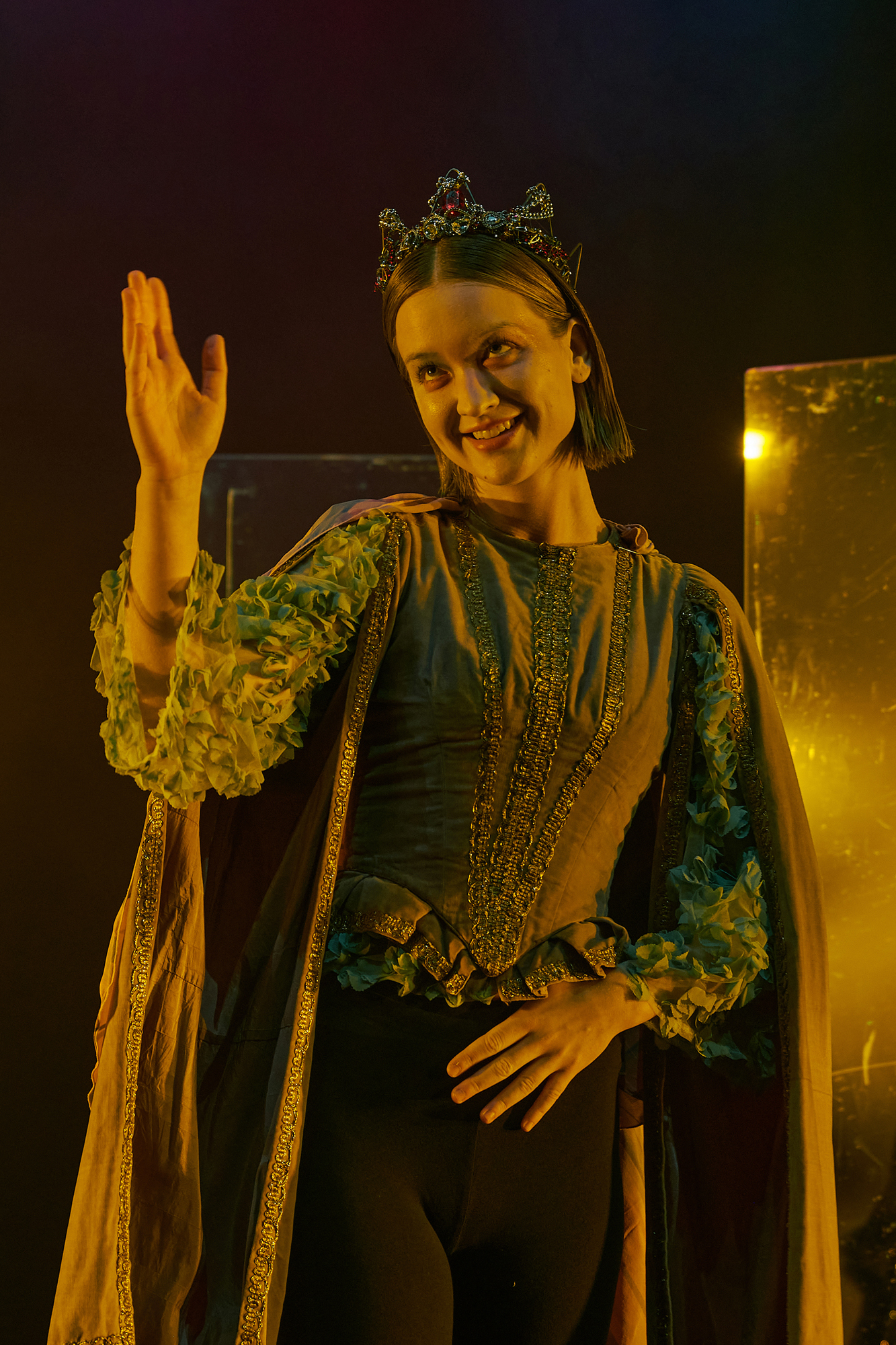 A photo of the character of Tintomara. She has a brunette page haircut and a crown. She is wearing a doublet with gold details. From the long sleeves hanging open, the sleeves of a sheer top with turqouise organza flowers peek out. Over her shoulders is a pink cape and on her lower half she wears black yoga pants. She is lit in golden yellow and is waving to someone out of frame. 