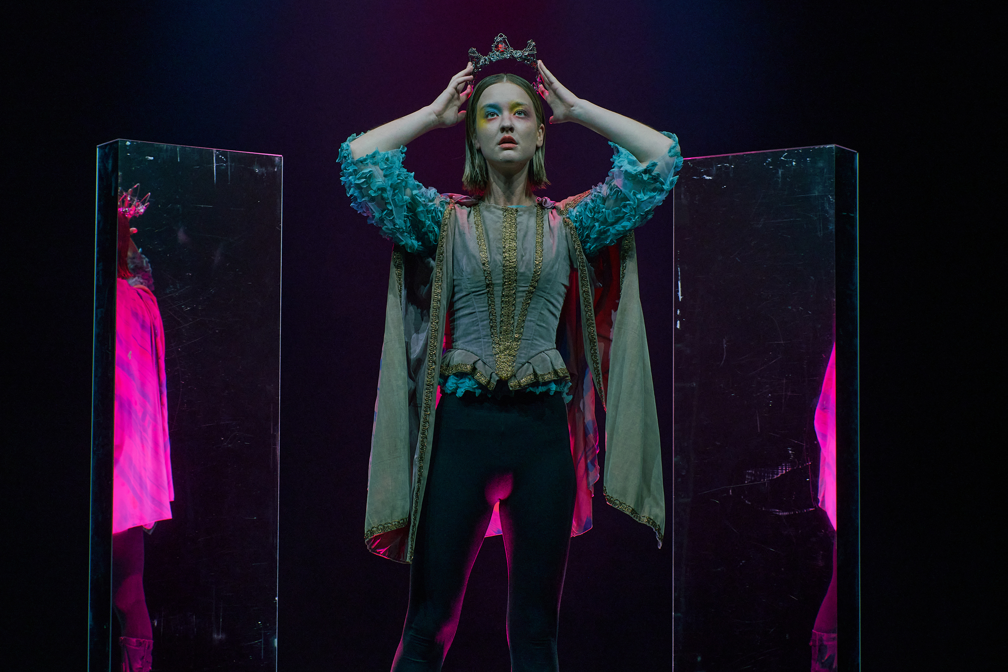 A photo of Tintomara. She is standing up looking into the distance, holding the crown over her head. Behind her are two mirrors, reflecting her cape.