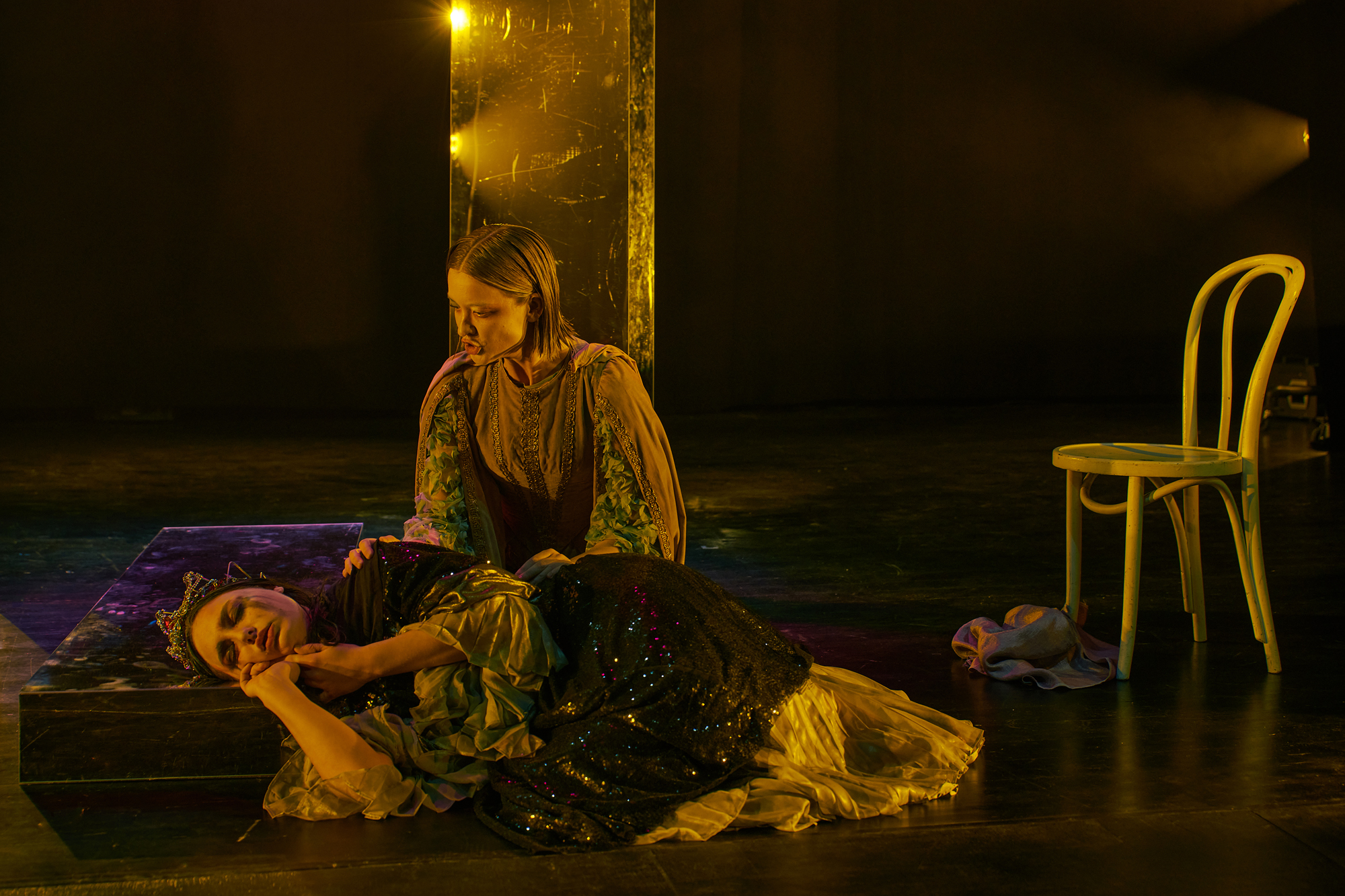 A photo of Clara and Tintomara. Clara is laying on the floor with her head resting on a laying down mirror. Tintomara is sitting behind her with her hand on her shoulder. Clara is wearing the crown, her eyes closed, while Tintomara sings to her. They are lit in gold, behind them are two more mirrors, standing up.