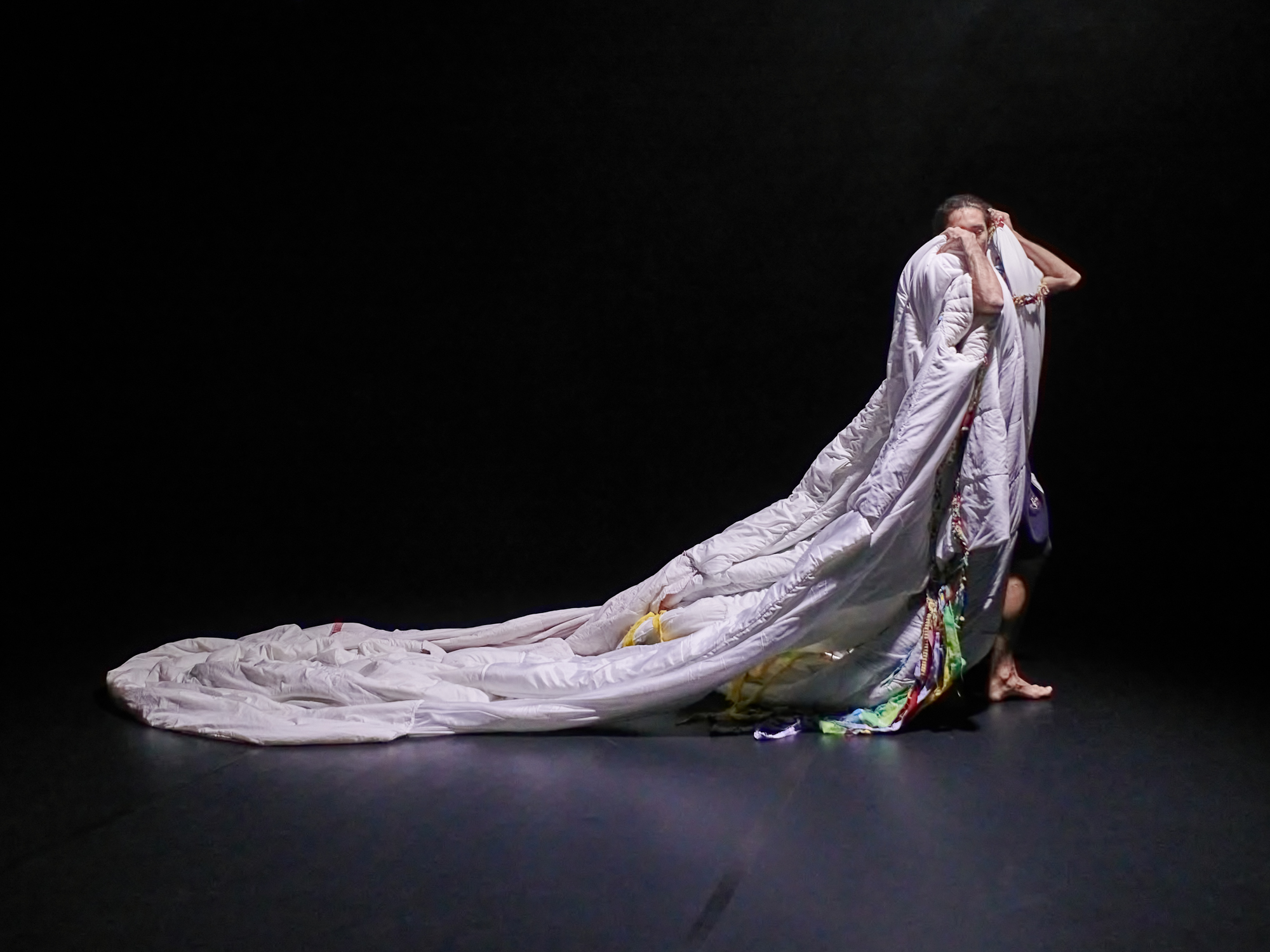 A photo of a dancer, mid-step, standing tall and wearing the a garment made out of duvets. It puffs over his shoulders and trails behind him like a cape. The trail is perhaps three meters.