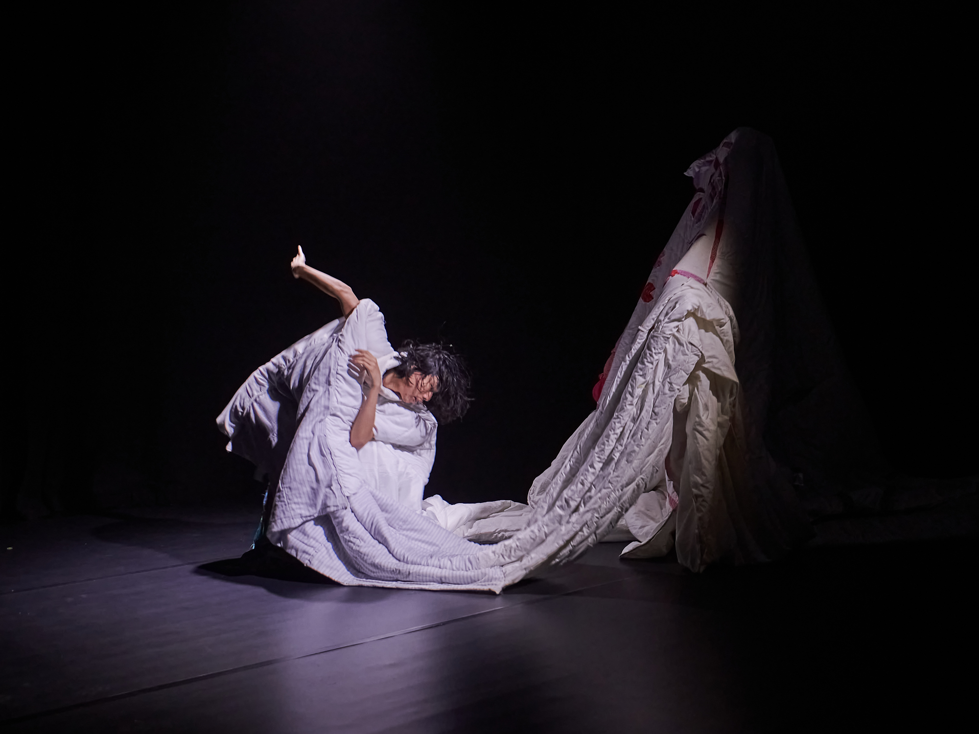 The same photo as the thumbnail. A photo of a dancer in a dark room. The dancer is crouched low to the ground, mid-movement, with one arm to his chest and the other outstretched behind him. He wears a bright white, unshapely garment, made out of several sewn together duvets. The garment drops to the ground in front of him before continuing up into a pillar-like shape into the dark.